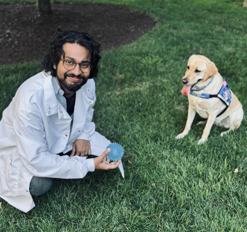 A photo of Dr. D'Souza in a white lab coat crouched in some grass holding a doggie toy. Across from Dr. D'Souza is a Golden Retriever with a harness on and it's tongue hanging out of its mouth.